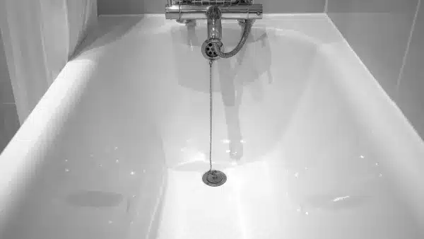 A white, empty bathtub with a silver metal drain plug and chain—ideal for easy drain cleaning; faucet and hose are visible on the tiled wall above.