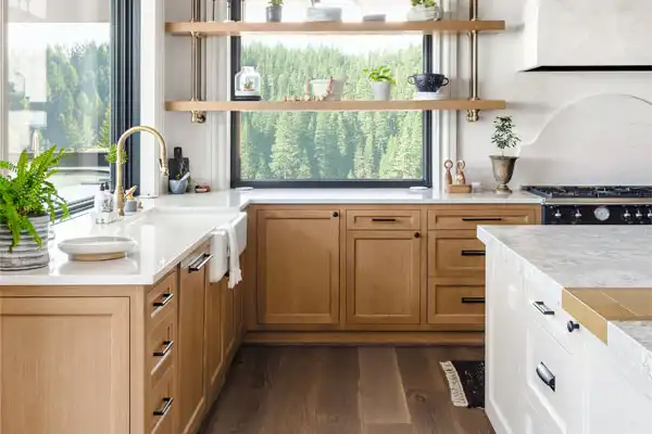 Modern kitchen with light wood cabinets, white countertops, open shelves with plants and dishes, large window with forest view, and gold faucet—perfect for those seeking reliable plumber repair or a trusted plumber near me.