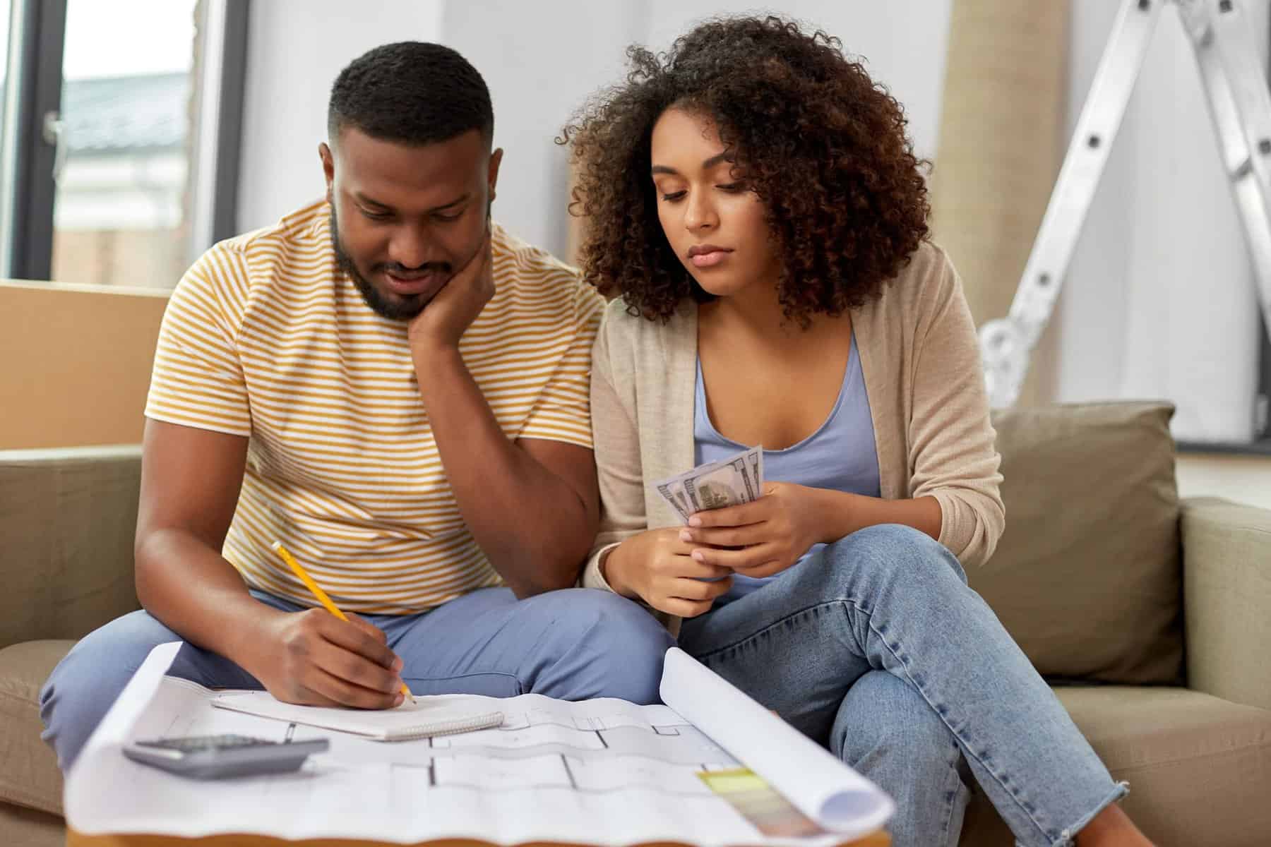 A man writes on a notepad while a woman next to him holds cash; a calculator, floor plans, and a DIY HVAC guide are on the table in front of them.
