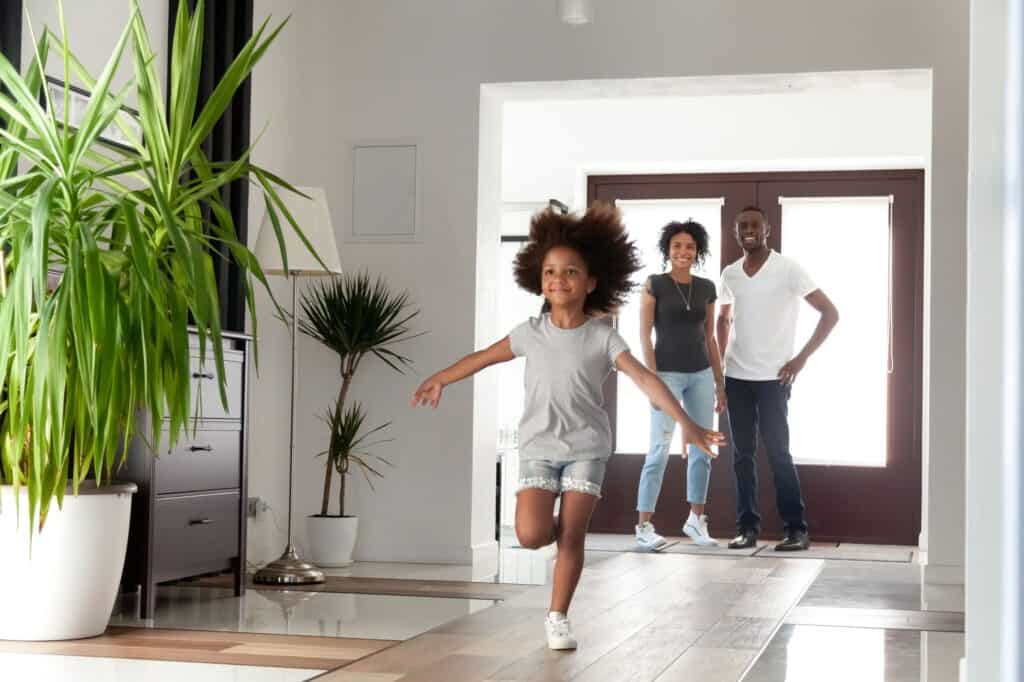 A young girl runs indoors with arms outstretched while two adults stand smiling by the doorway in a modern, well-lit home with potted plants and the best air conditioning keeping everyone cool and comfortable.