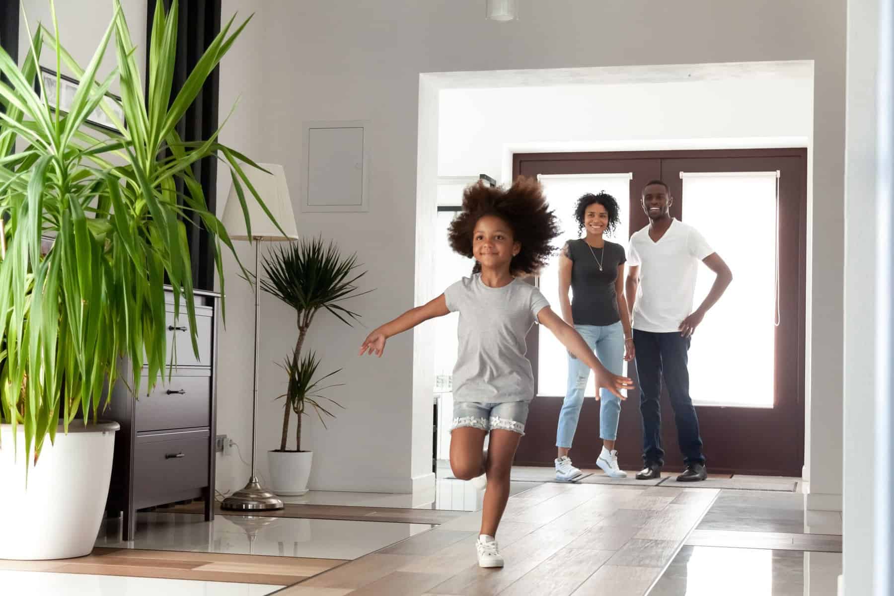 A young girl runs indoors with arms outstretched while two adults stand smiling by the doorway in a modern, well-lit home with potted plants and the best air conditioning keeping everyone cool and comfortable.