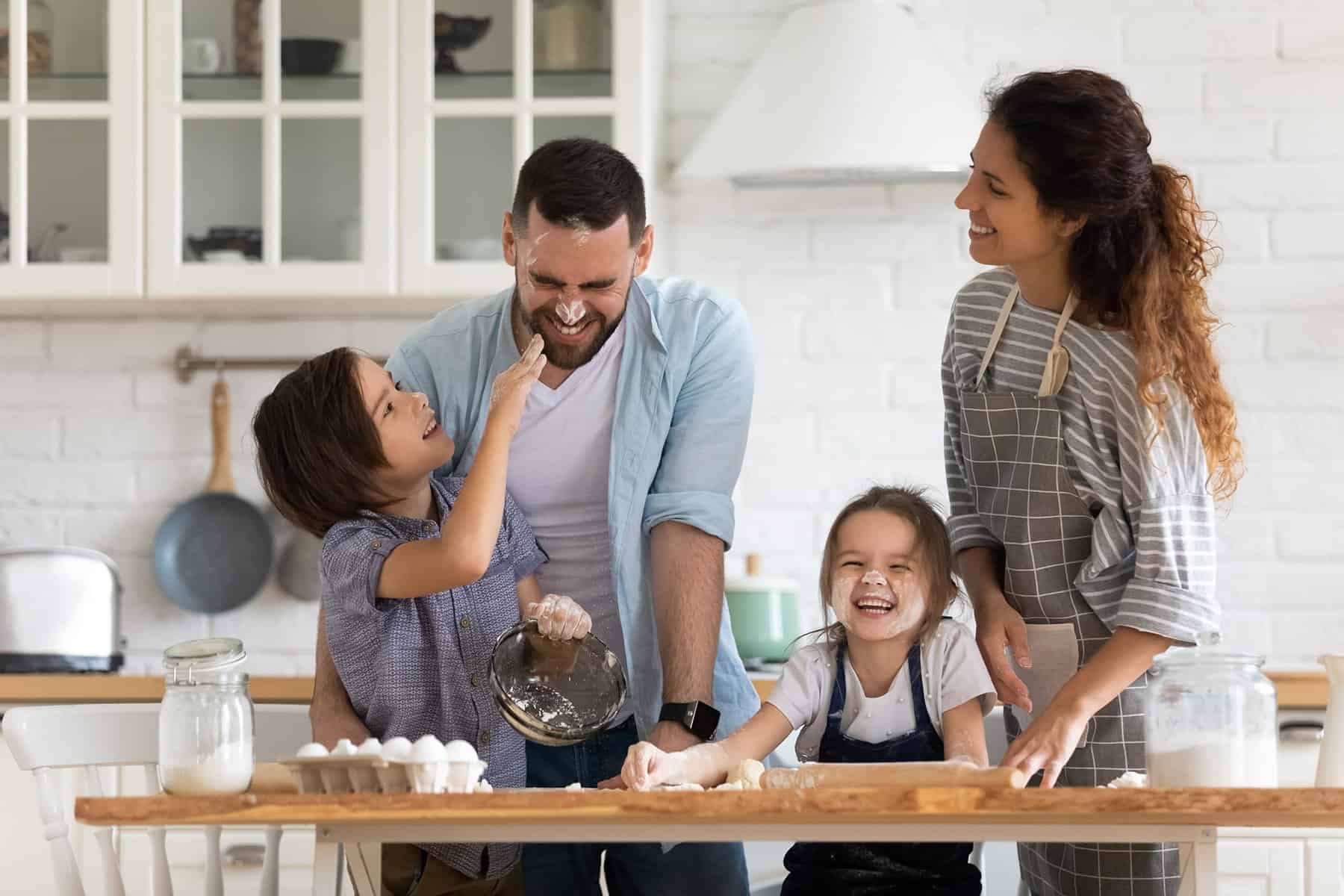 A family of four smiles and laughs together while baking in a bright kitchen, unaware that copper pipes beneath the floor might need repair or replace services due to a small pinhole leak. Flour and baking ingredients are visible on the wooden table.