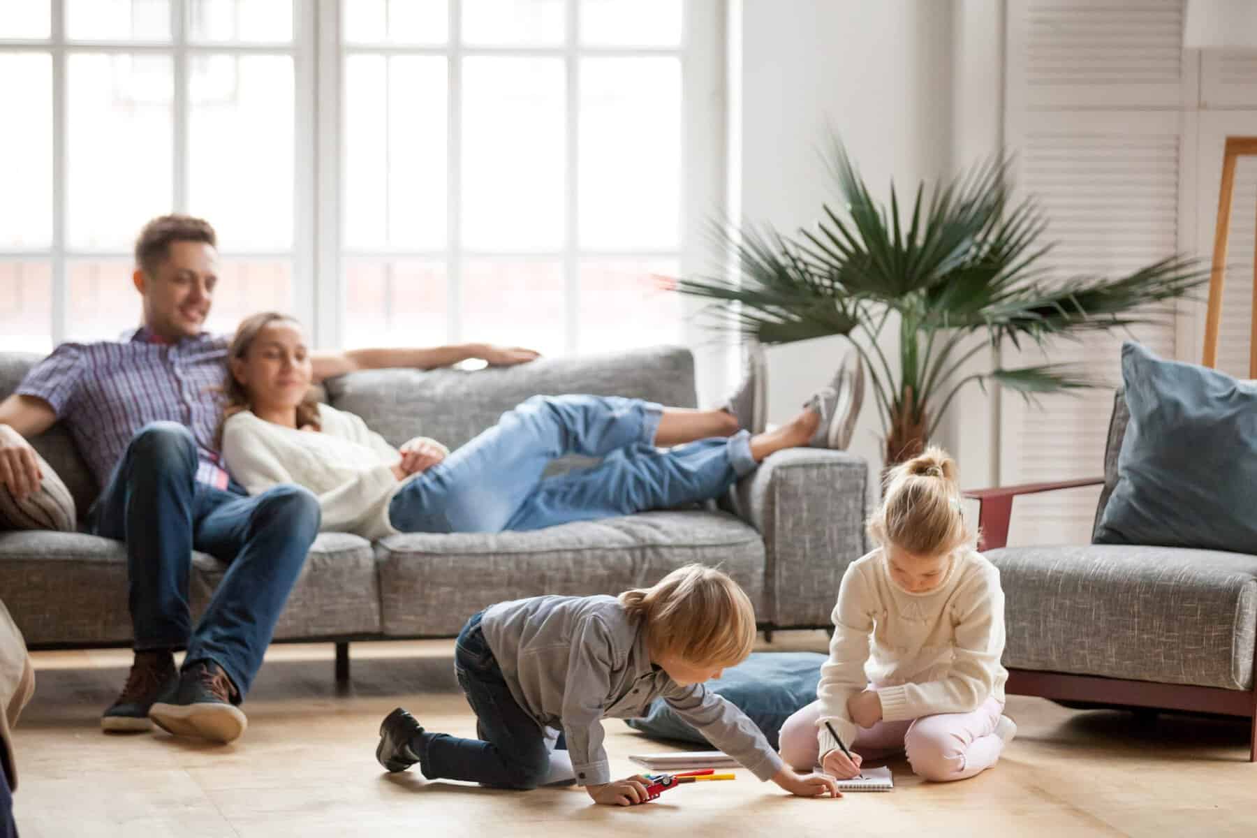 Two children draw on the floor with colored pencils while two adults relax on a gray couch in a bright living room, enjoying the health benefits of home humidifiers and their close family relationship.