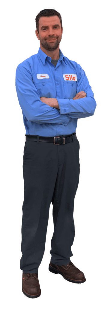 A man in a blue button-up shirt with a "Sila" logo, dark pants, and brown shoes stands with arms crossed, facing forward and smiling.