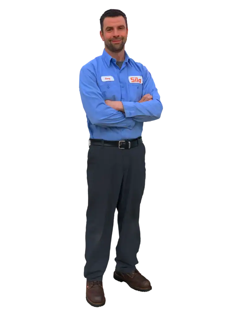 A man wearing a blue work shirt with a "Sila" logo and name tag, dark pants, and brown shoes stands with arms crossed, smiling.