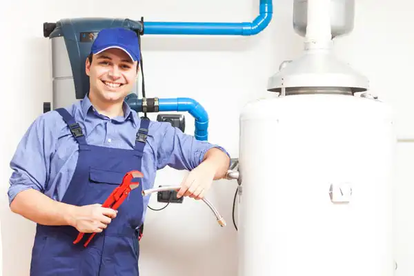 A plumber in blue overalls and cap holds tools, standing next to a white water heater with visible piping on the wall behind him, ready for boiler maintenance in Beltsville, MD.