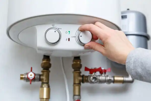 A person adjusts the temperature dial on a wall-mounted water heater after a recent boiler installation in Beltsville, MD, with visible pipes and valves below the unit.