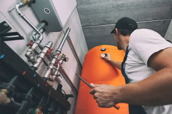 A technician inspects and adjusts controls on an orange hot water tank in a utility room with exposed pipes and valves during a heating repair in Beltsville MD.