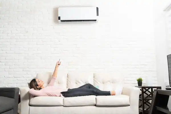 A woman relaxes on a sofa in her bright Beltsville living room, pointing a remote at her wall-mounted mini-split installation for efficient cooling in MD.