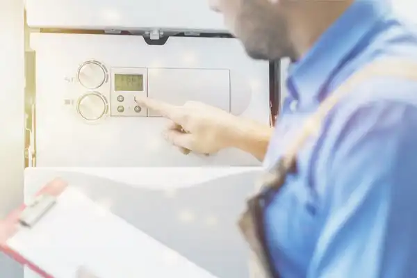A person in work attire adjusts the controls on a wall-mounted boiler or HVAC unit in Beltsville, MD, while holding a clipboard—possibly overseeing a tankless water heater installation.