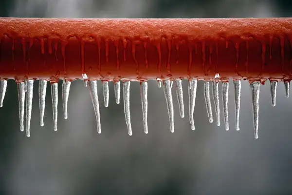 A red metal pipe with multiple icicles hanging from its underside against a blurred gray background, highlighting the need for frozen pipe repair in Beltsville MD.