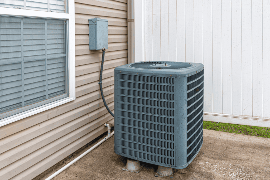 A central air conditioning unit is installed outside a house next to a beige exterior wall, near a window and an electrical box, showcasing the benefits of central air for efficient, whole-home cooling.