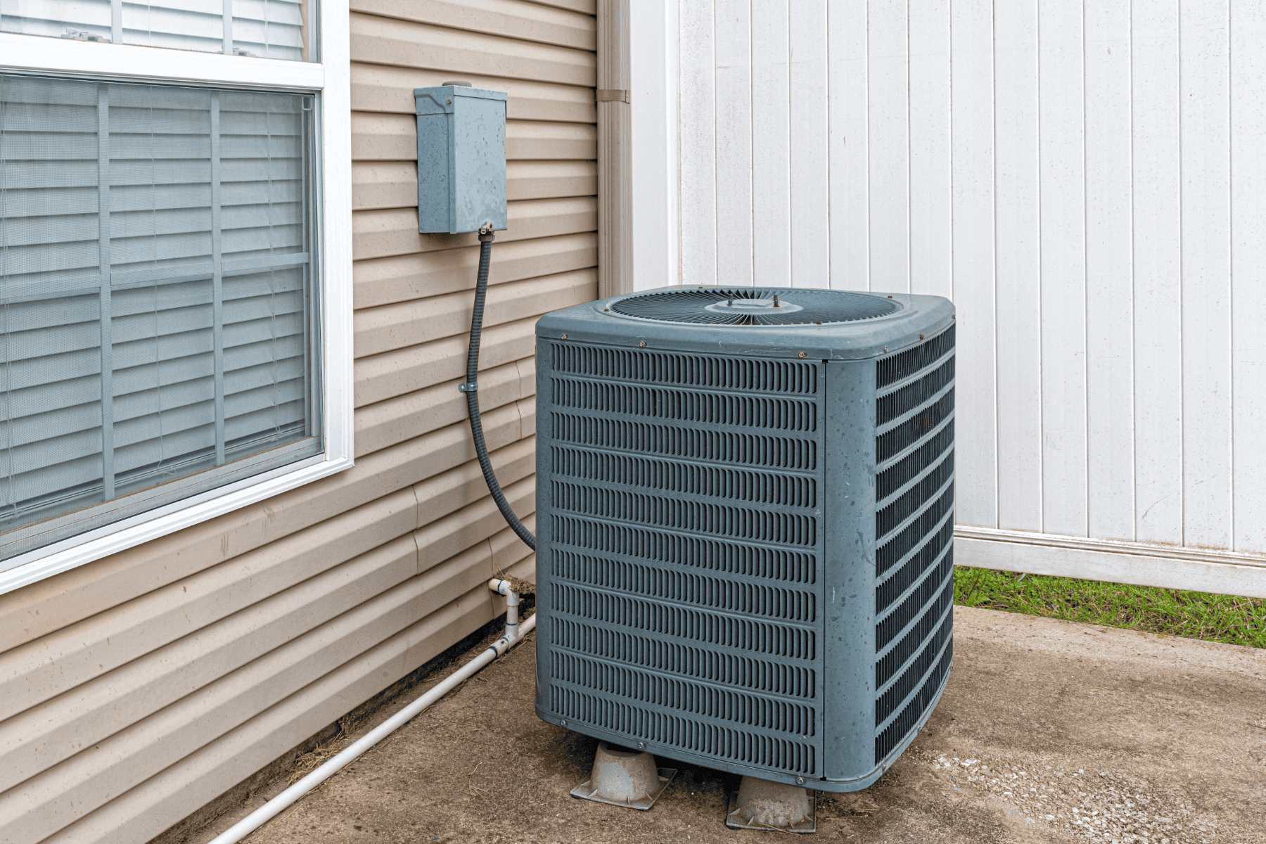 A central air conditioning unit is installed outside a house next to a beige exterior wall, near a window and an electrical box, showcasing the benefits of central air for efficient, whole-home cooling.