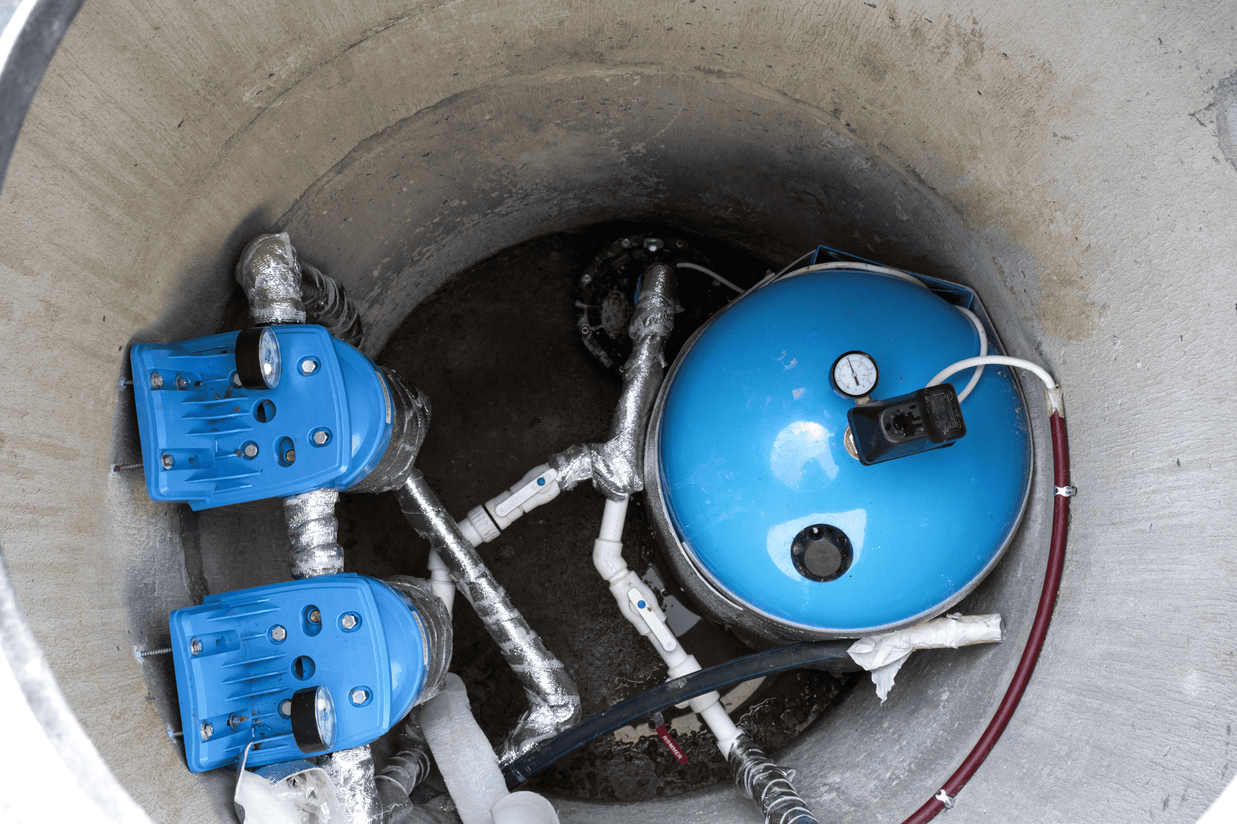 A concrete well contains a blue pressure tank, gauge, pipes, and two blue water pump controllers with foil insulation wrapping on some pipes.