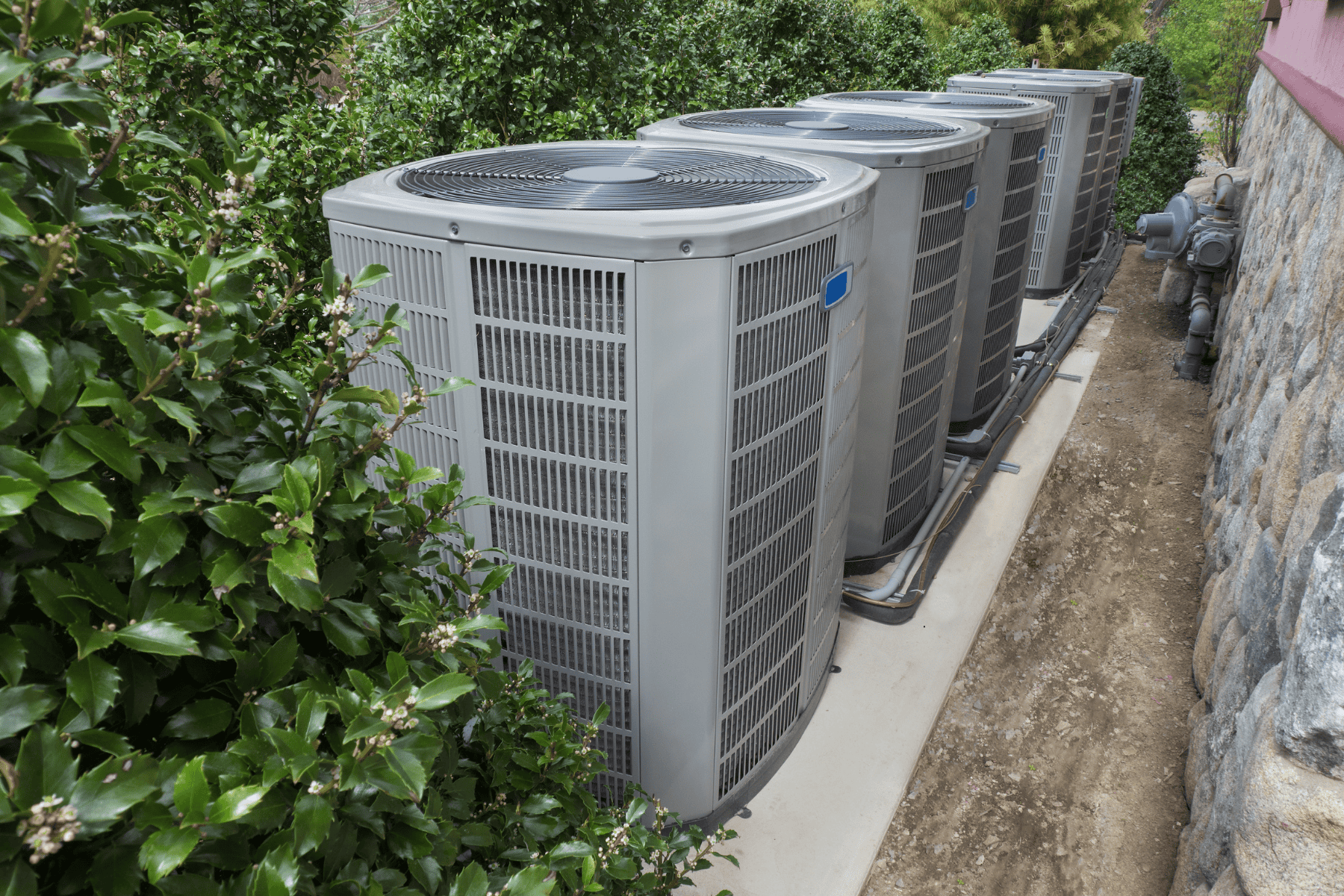 Four large outdoor air conditioning units are positioned in a row on a concrete slab next to a building, with greenery on one side and a stone wall on the other—a modern solution often chosen for older homes or historic homes seeking efficient high velocity air conditioning without compromising character.