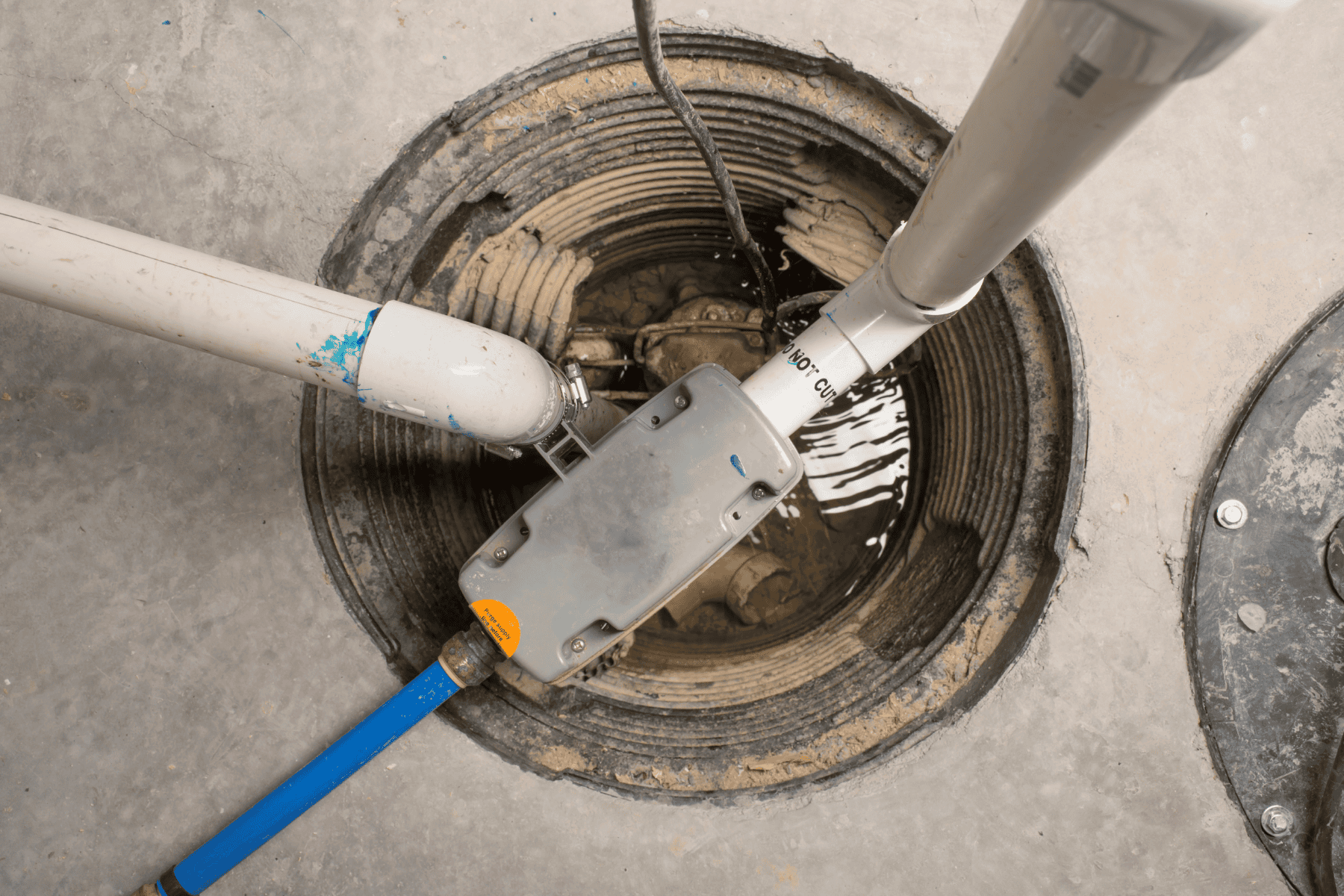 A sump pump with attached pipes, a sump pump check valve, and electrical wiring is installed in a circular pit filled with some water, set in a concrete floor.