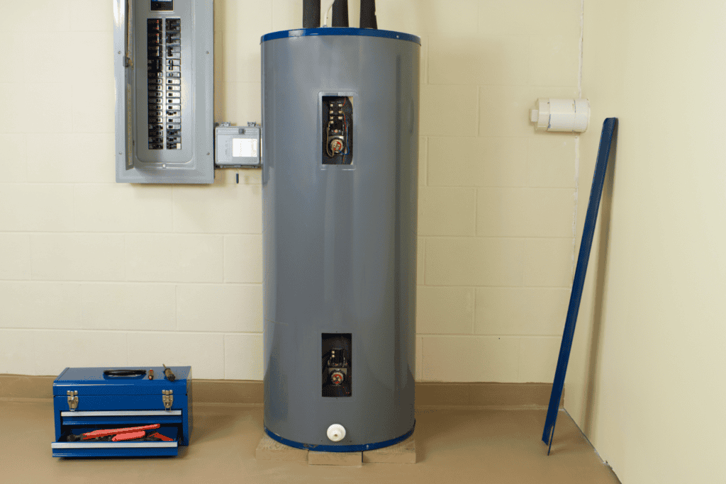 Electric water heater with open access panels positioned next to a breaker panel and a blue metal toolbox, showing the pressure relief valve.