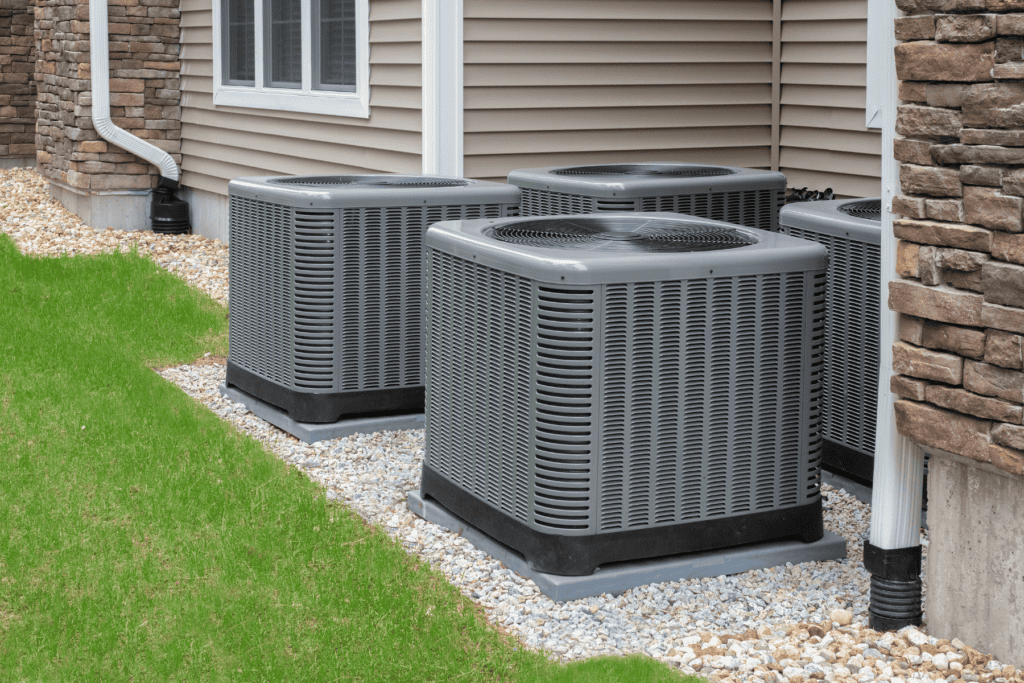 Three grey central air conditioning units, including a failing heat pump, placed on gravel next to a house exterior.